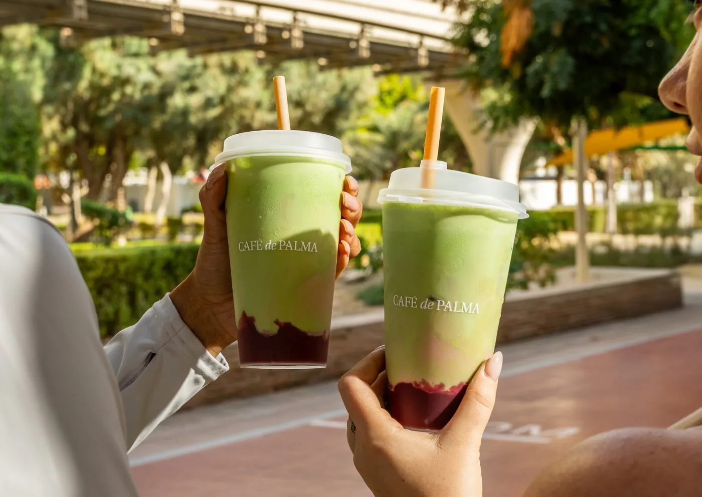 Two smoothies in Café de Palma cups, held up outdoors in the sun